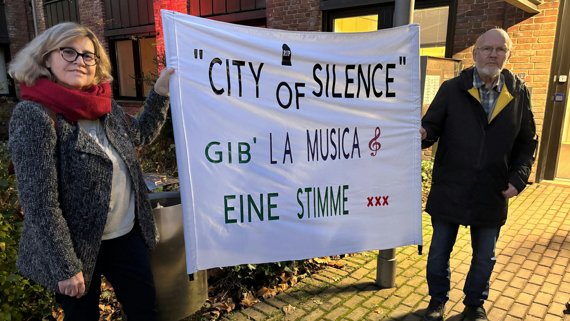 Das Foto zeigt zwei Menschen, die ein Plakat mit der Aufschrift „City of Silence Gib La Musica eine Stimme“ beschriftet ist.