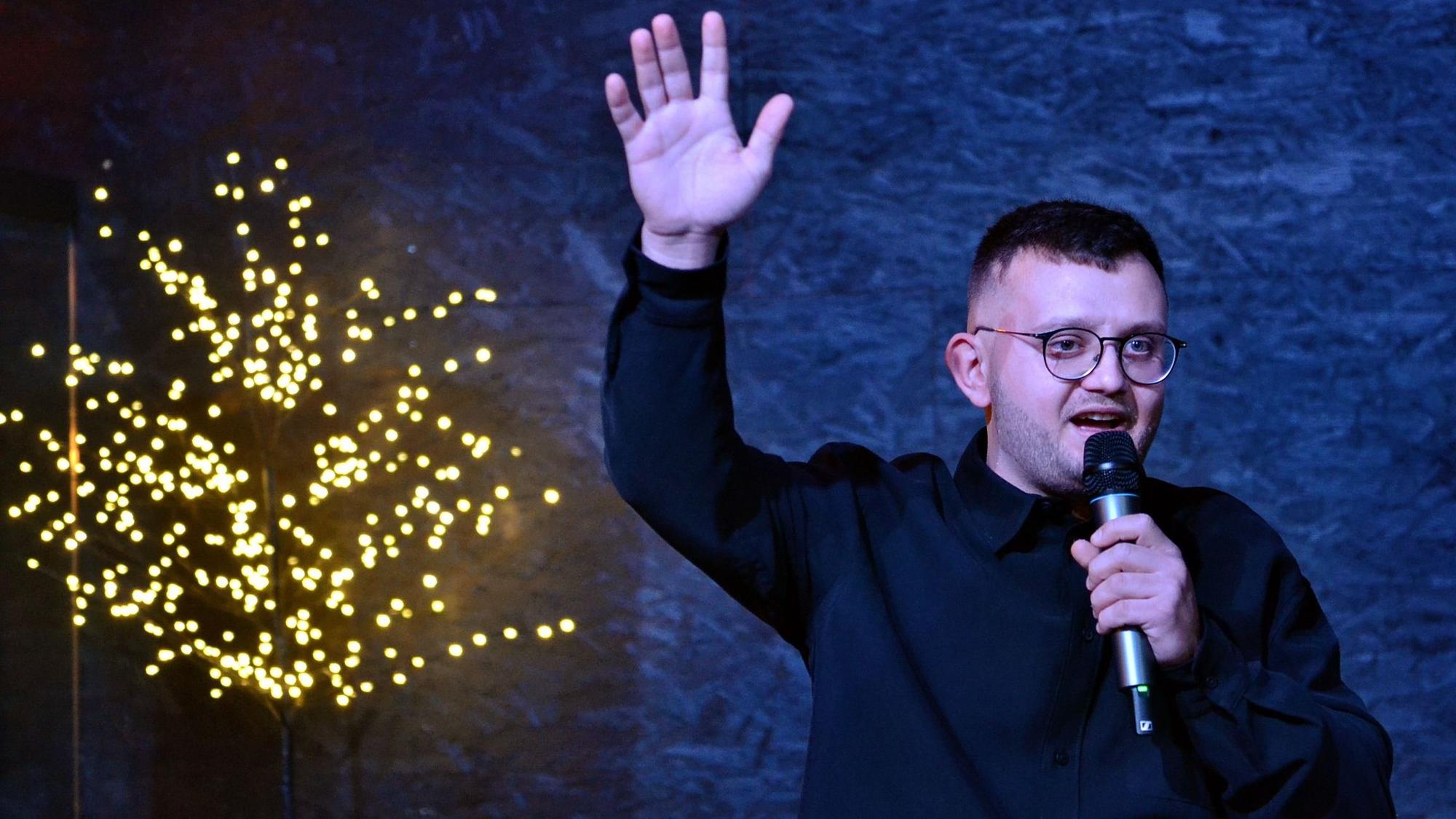 Veranstalter Mykola Pavlovtsev hat ein Mikrofon in der einen Hand, die andere hat er erhoben. Im Hintergrund sieht man einen mit Lichterketten behängten Baum.