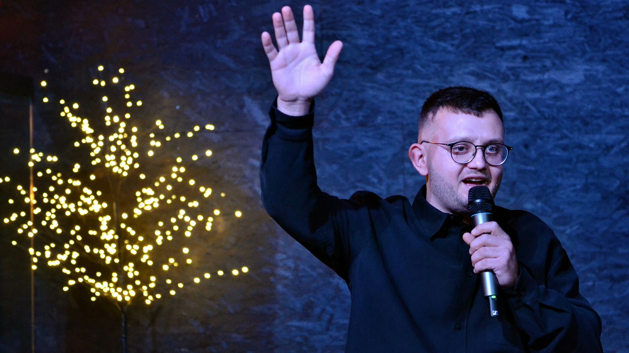 Veranstalter Mykola Pavlovtsev hat ein Mikrofon in der einen Hand, die andere hat er erhoben. Im Hintergrund sieht man einen mit Lichterketten behängten Baum.