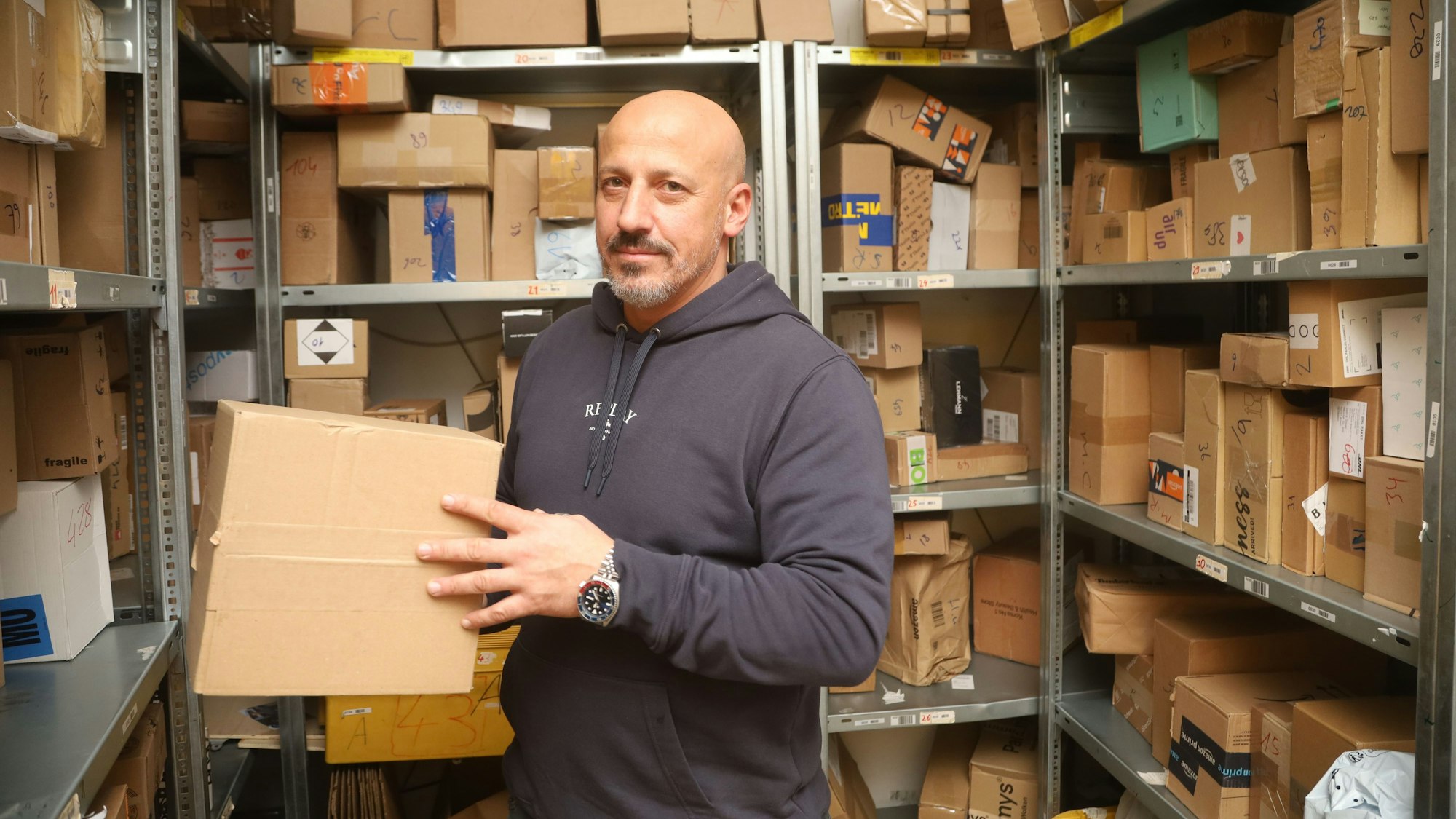 Ein Mann hält ein Paket in der Hand. Hinter ihm befinden sich Pakete in Regalen.