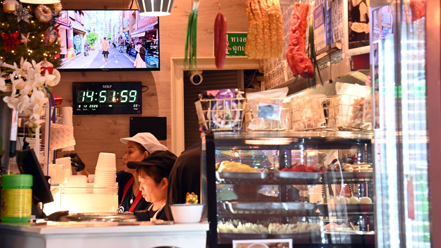 Blick in das Lokal Thai Street Food: Vitrinen mit Essen, Lebensmittel hängen von der Decke, Blumen und Weihnachtsschmuck stehen auf der Theke, dahinter arbeiten 2 Menschen konzentriert.