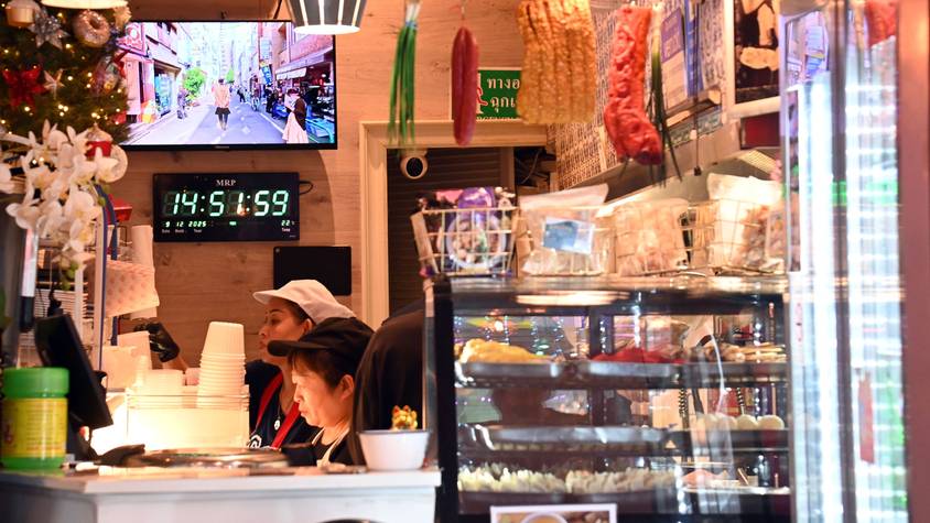 Blick in das Lokal Thai Street Food: Vitrinen mit Essen, Lebensmittel hängen von der Decke, Blumen und Weihnachtsschmuck stehen auf der Theke, dahinter arbeiten 2 Menschen konzentriert.