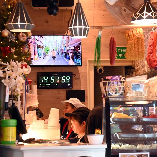 Blick in das Lokal Thai Street Food: Vitrinen mit Essen, Lebensmittel hängen von der Decke, Blumen und Weihnachtsschmuck stehen auf der Theke, dahinter arbeiten 2 Menschen konzentriert.