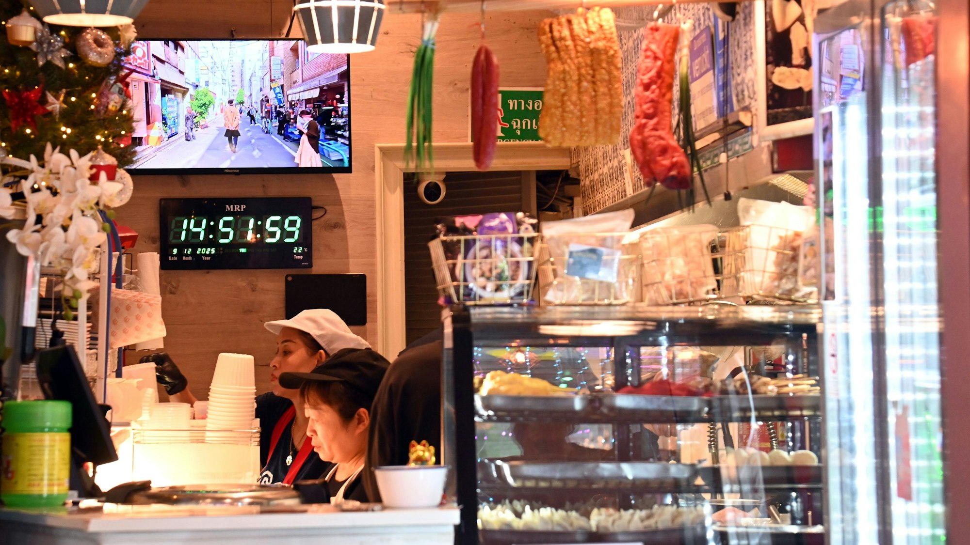 Blick in das Lokal Thai Street Food: Vitrinen mit Essen, Lebensmittel hängen von der Decke, Blumen und Weihnachtsschmuck stehen auf der Theke, dahinter arbeiten 2 Menschen konzentriert.