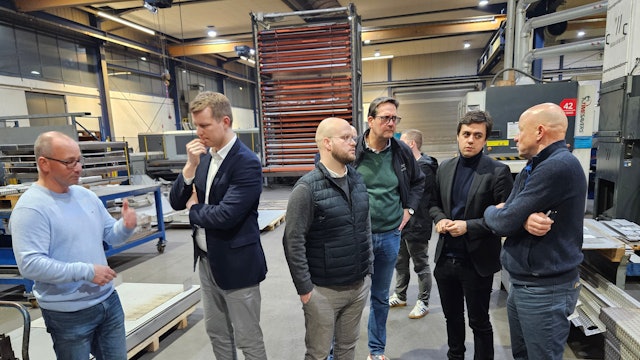 Sechs Männer stehen in einer Werkhalle und sprechen miteinander.