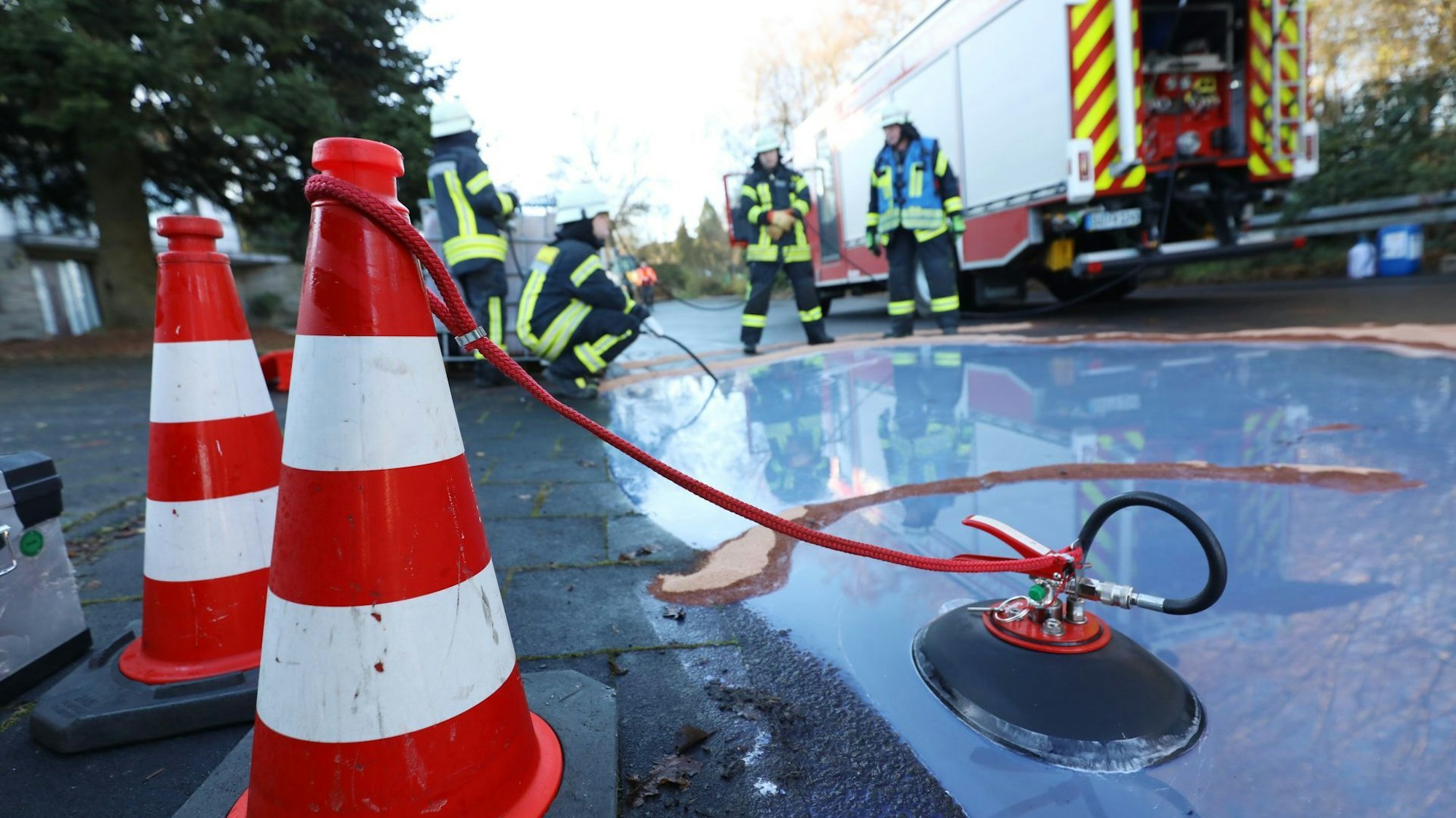 Ein kleiner „See“ mit Löschwasser am Straßenrand, im Hintergrund vier Feuerwehrleute.