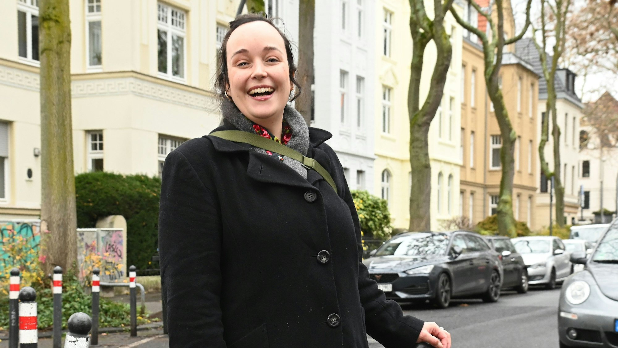 Eine junge Frau steht an einem Straßenpoller, hinter ihr sind schöne Altbauten in der Ottostraße in Köln zu sehen.