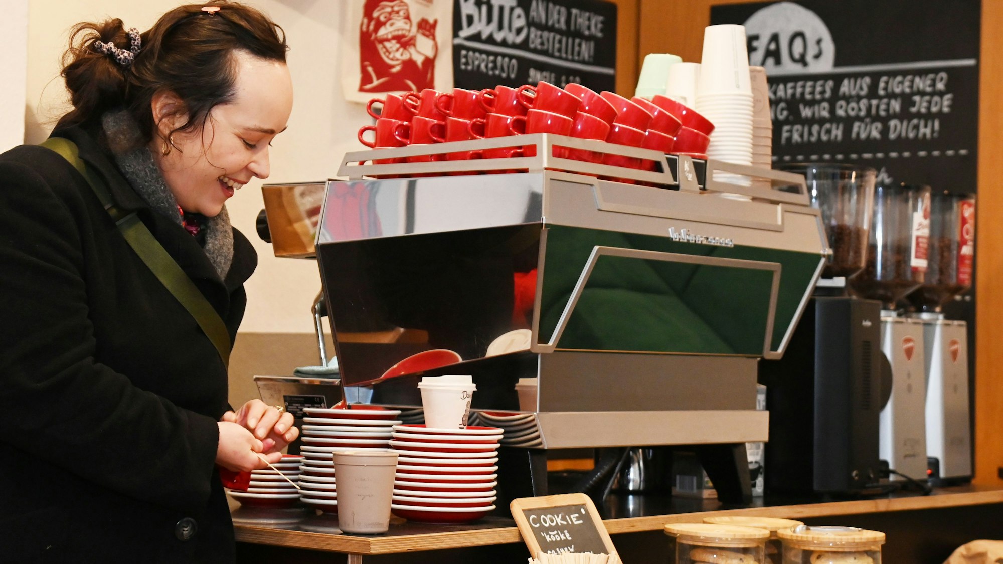 Eine Frau mit dunklem Haar und dunklem Mantel steht an einer Theke, auf der eine Kaffeemaschine steht.