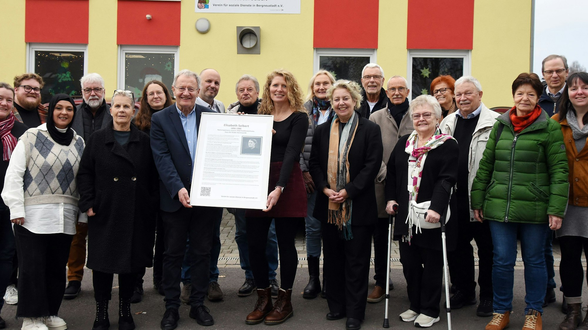 Blick auf eine Gruppe von Menschen, die vor einem Kindergarten eine Namenstafel präsentieren.