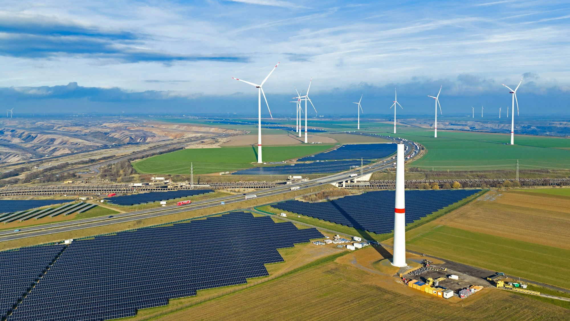 Zu sehen sind Solarmodule, fertige Windräder und im Vordergrund ein im Bau befindliches Windrad.