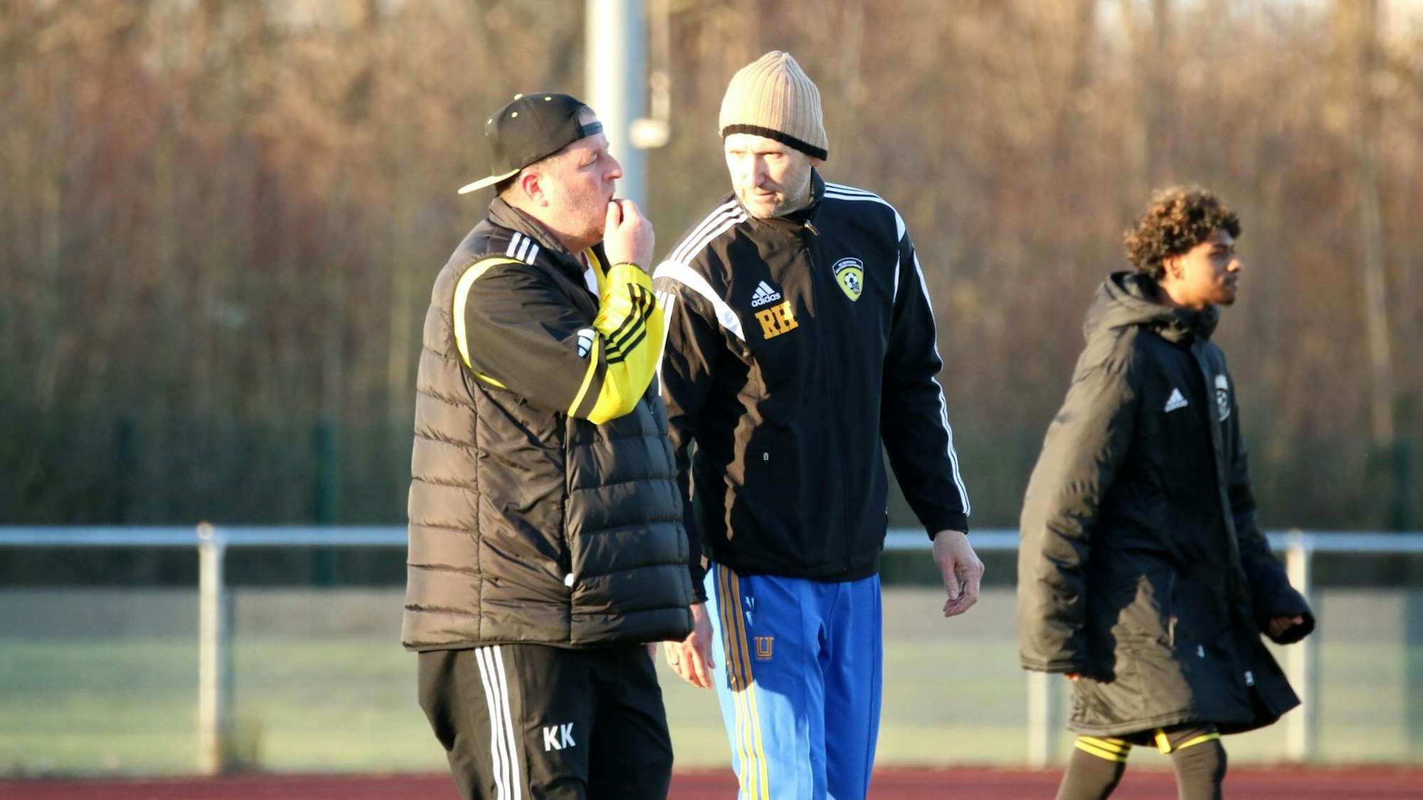 Karsten Kochems kratzt sich am Kinn, er trägt eine Baseballkappe falsch herum. Ralph Hoppen spricht mit ihm, er trägt eine Mütze.
