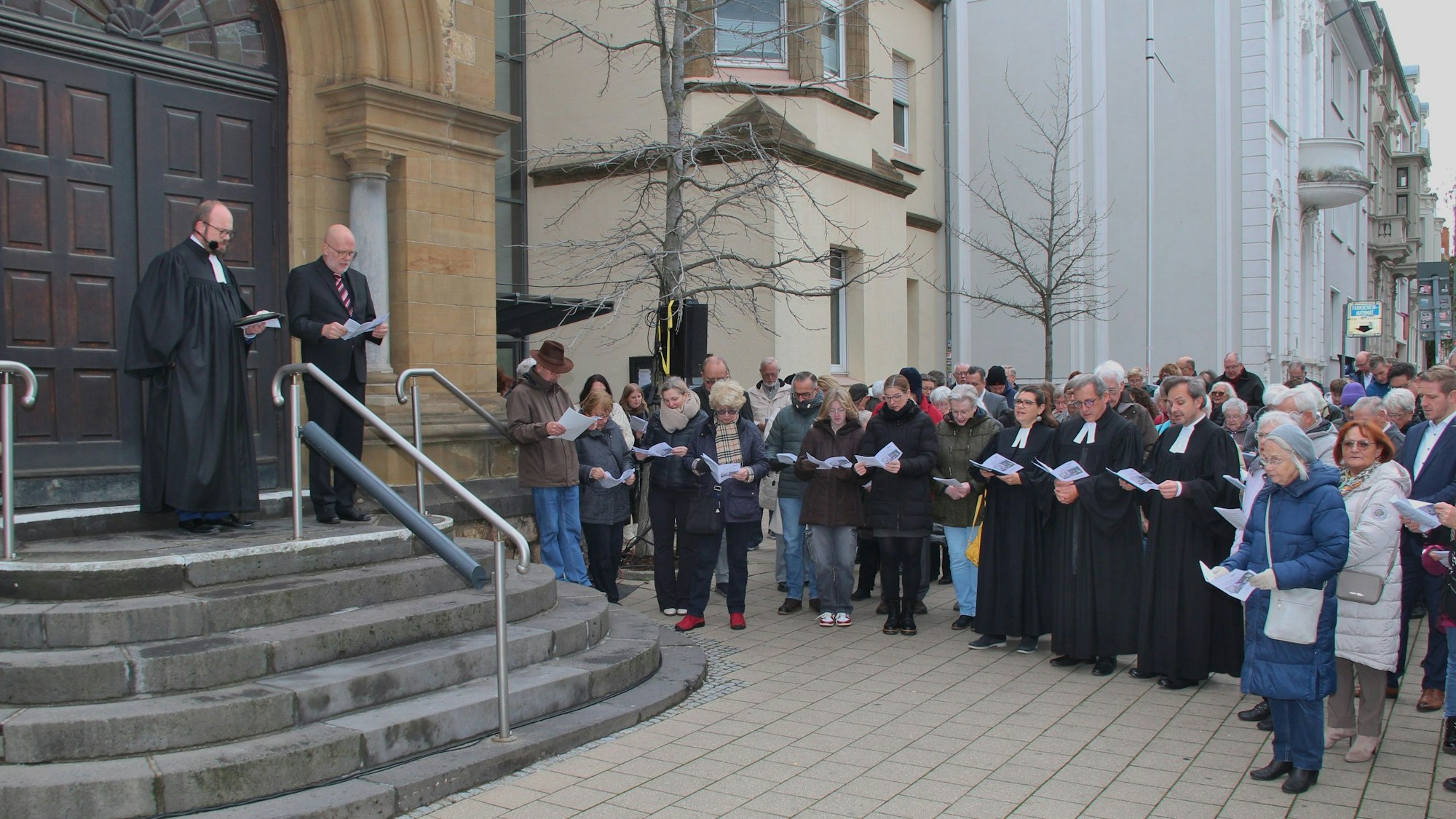 Gregor Weichsel und Michael Bork stehen am oberen Ende der Treppe vor der Kirchentür. Die Gemeidemitglieder haben Texthefte in der Hand.