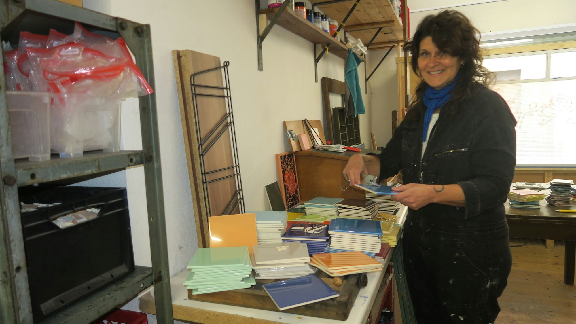 Eine Frau mit dunkem Haar steht an einem Tisch, auf dem Fliesen in unterschiedlichen Farben liegen.