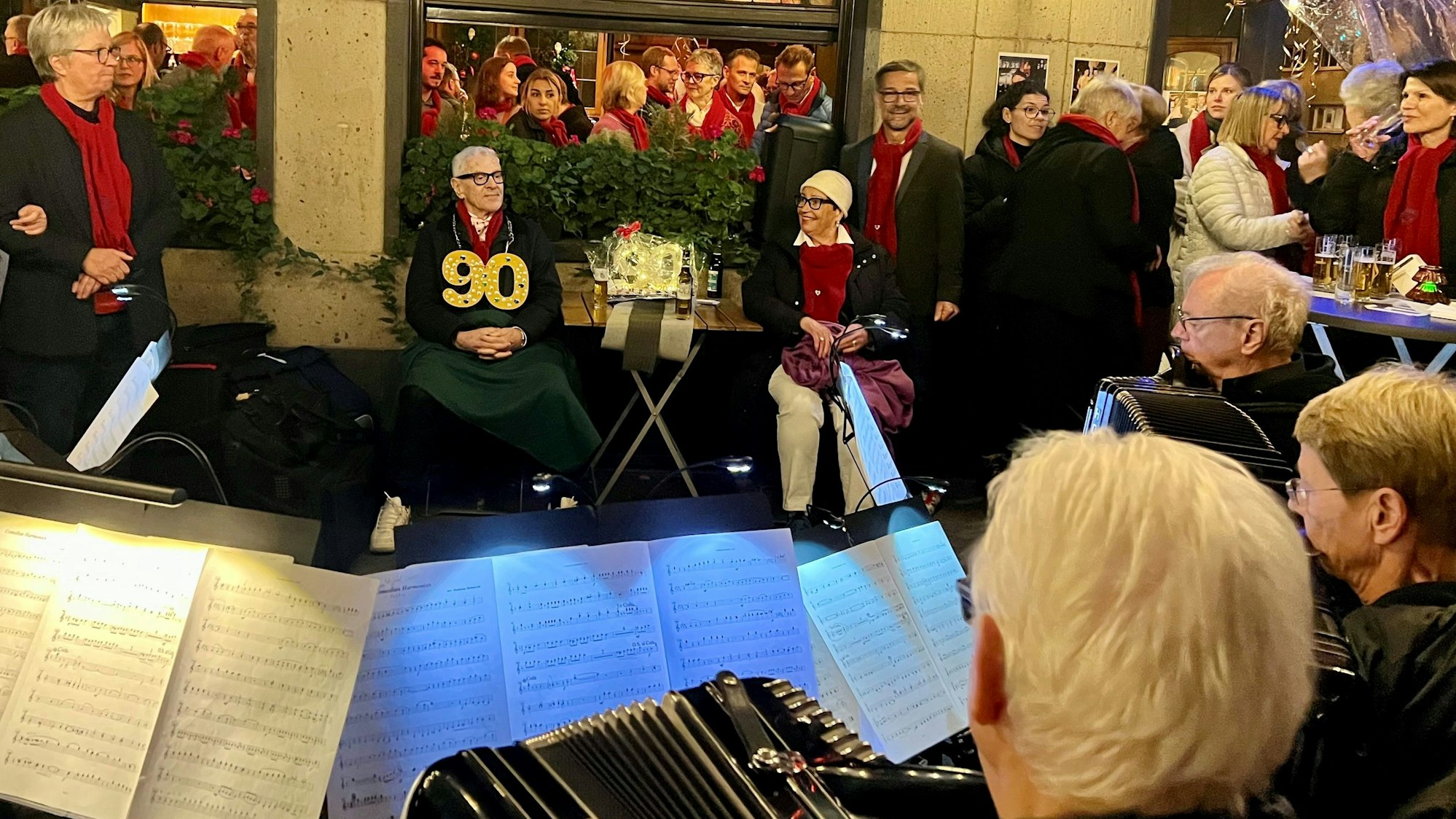 Vor einer Kneipe sitzt ein Paar an einem Tisch, auf dem Bürgersteig davor hat ein Akkordeon-Orchester Aufstellung genommen.