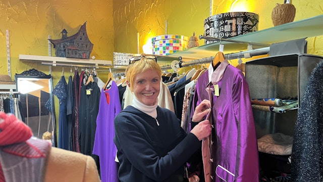 Eine ältere Frau mit kurzem blonden Haar steht in einem Bekleidungsgeschäft und knöpft eine lilafarbene Jacke zu.
