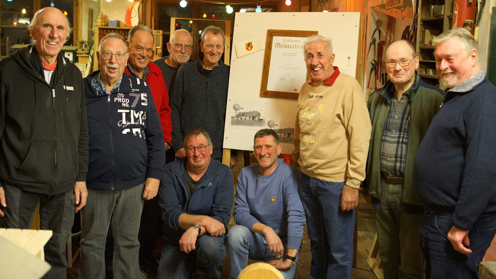 Schreiner Hans-Peter Lückerath steht vor dem gerahmten goldenen Meisterbrief. Er ist umringt von ehemaligen Gesellen.