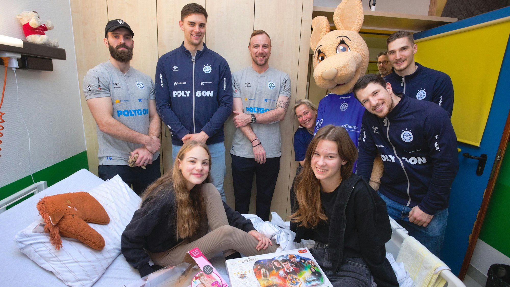 Zwei Mädchen sitzen auf dem Bett im Krankenhauszimmer, während die fünf Handball-Profis und das Maskottchen daneben stehen.