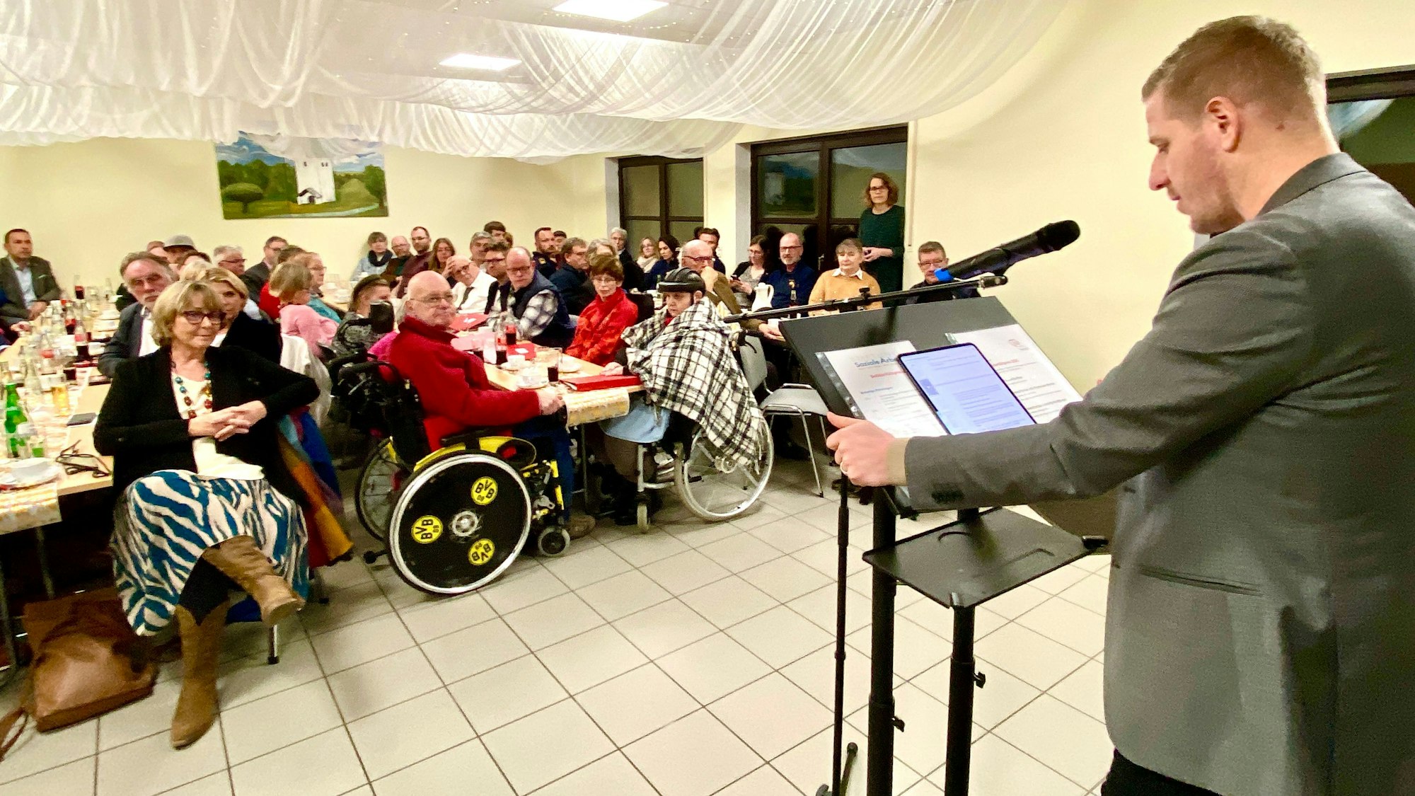 Der Landrat steht am Rednerpult, in dem Raum sitzen viele Menschen und hören ihm zu.