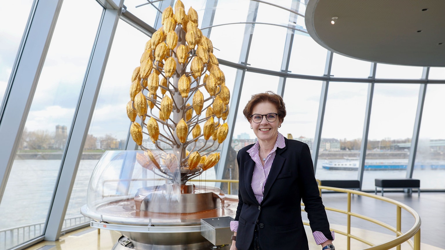 Annette Imhoff, Chefin des Kölner Schokoladenmuseums, steht vor einem stilisierte Kakao-Baum in Gold und lächelt in die Kamera.