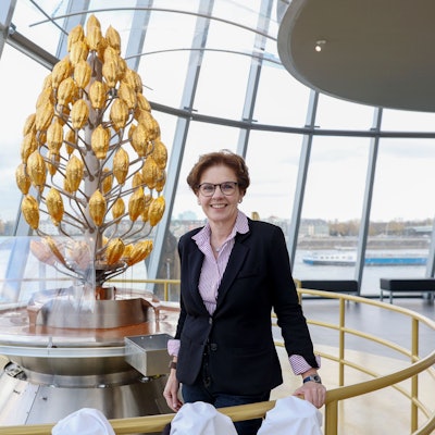 Annette Imhoff, Chefin des Kölner Schokoladenmuseums, steht vor einem stilisierte Kakao-Baum in Gold und lächelt in die Kamera.