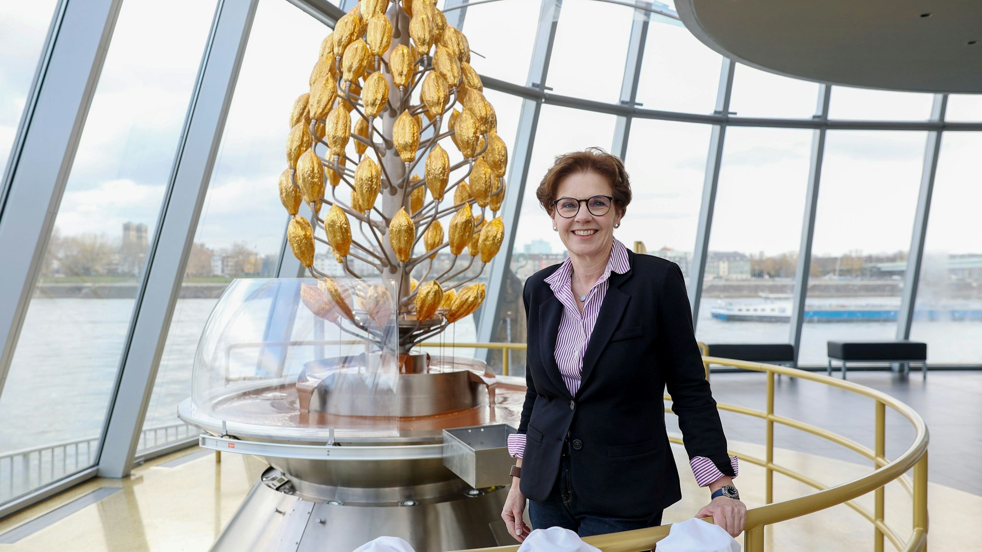 Annette Imhoff, Chefin des Kölner Schokoladenmuseums, steht vor einem stilisierte Kakao-Baum in Gold und lächelt in die Kamera.
