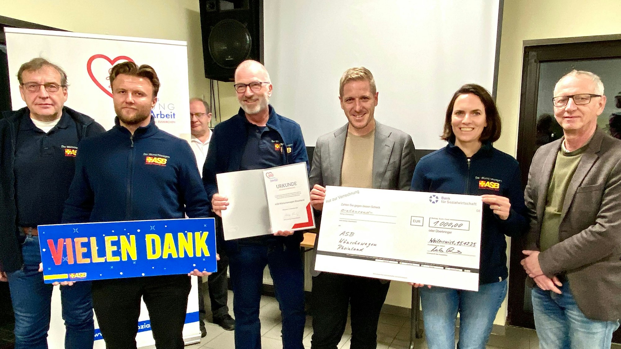 Mitglieder des Wünschewagen-Teams freuen sich über den symbolischen Riesen-Scheck, den sie überreicht bekamen. Zwei halten ein Schild mit den Worten „Vielen Dank“ in den Händen.