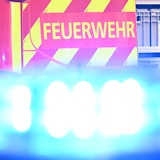 Ein Fahrzeug der Feuerwehr steht bei einem Einsatz auf einer Straße, während im Vordergrund das Blaulicht eines Polizeifahrzeugs zu sehen ist.