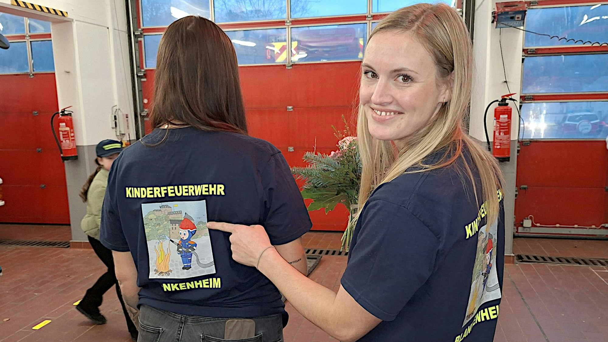 Kerstin Heller zeigt mit dem Finger auf den Rücken von Isabell Engel, deren T-Shirt mit dem Logo der Kinderfeuerwehr Blankenheim bedruckt ist.