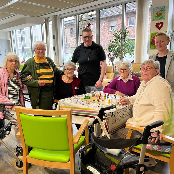 Fünf Seniorinnen und zwei Pflegekräfte stehen oder sitzen in einem Gemeinschaftsraum einer Tagespflege.