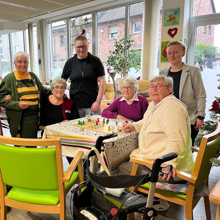 Fünf Seniorinnen und zwei Pflegekräfte stehen oder sitzen in einem Gemeinschaftsraum einer Tagespflege.