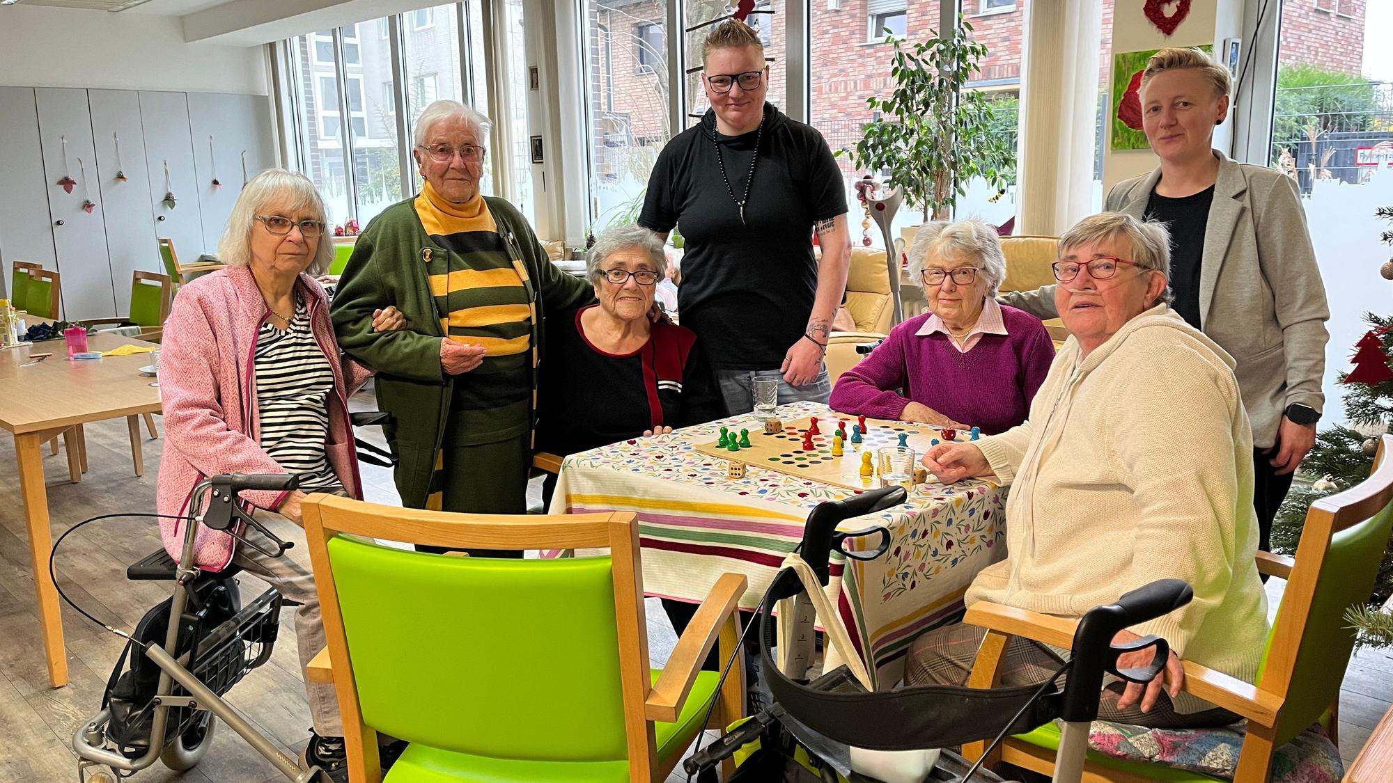 Fünf Seniorinnen und zwei Pflegekräfte stehen oder sitzen in einem Gemeinschaftsraum einer Tagespflege.