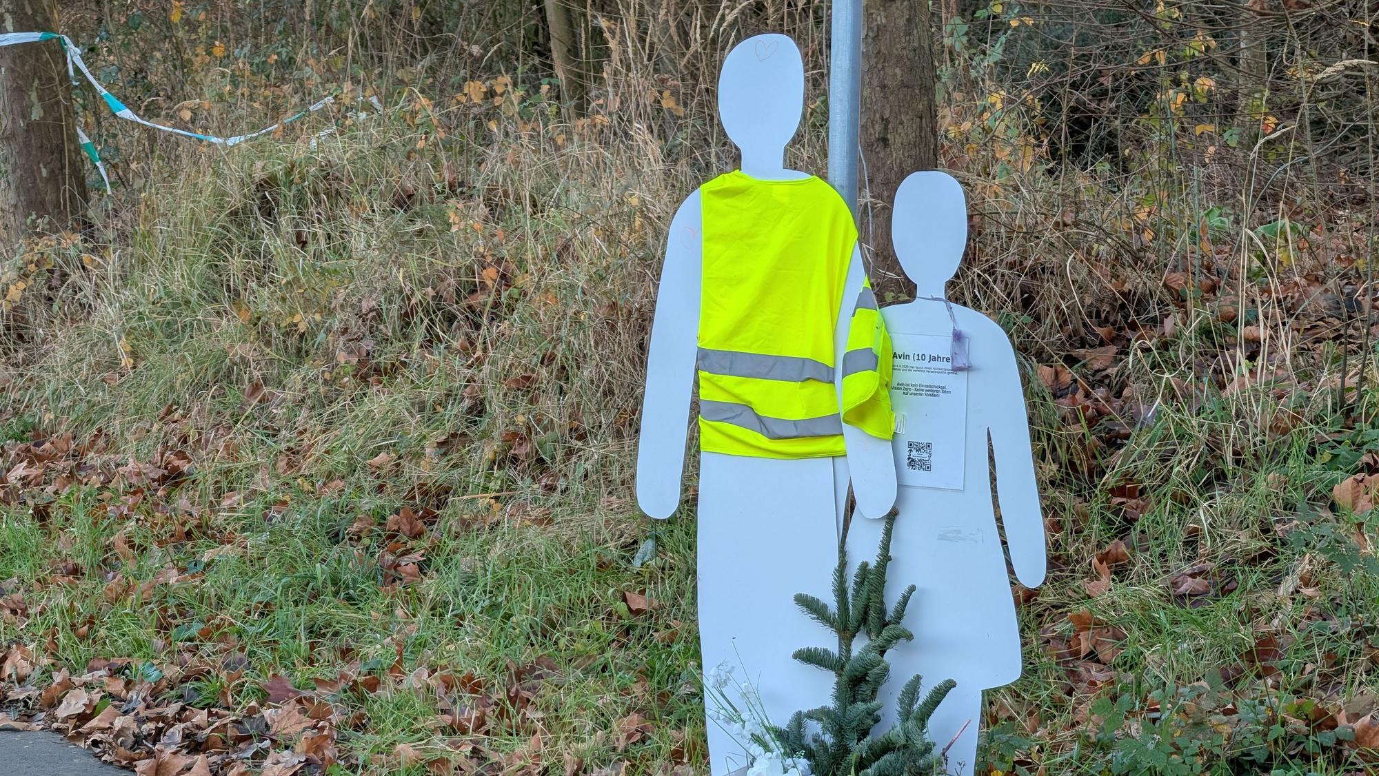 Das Foto zeigt zwei Holzfiguren am Straßenrand, eine davon trägt eine Sicherheitsweste.