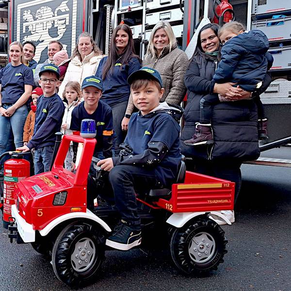 Ein Junge sitzt auf einem Mini-Feuerwehrfahrzeug. Neben ihm stehen weitere Kinder, hinter ihnen einige Erwachsene.