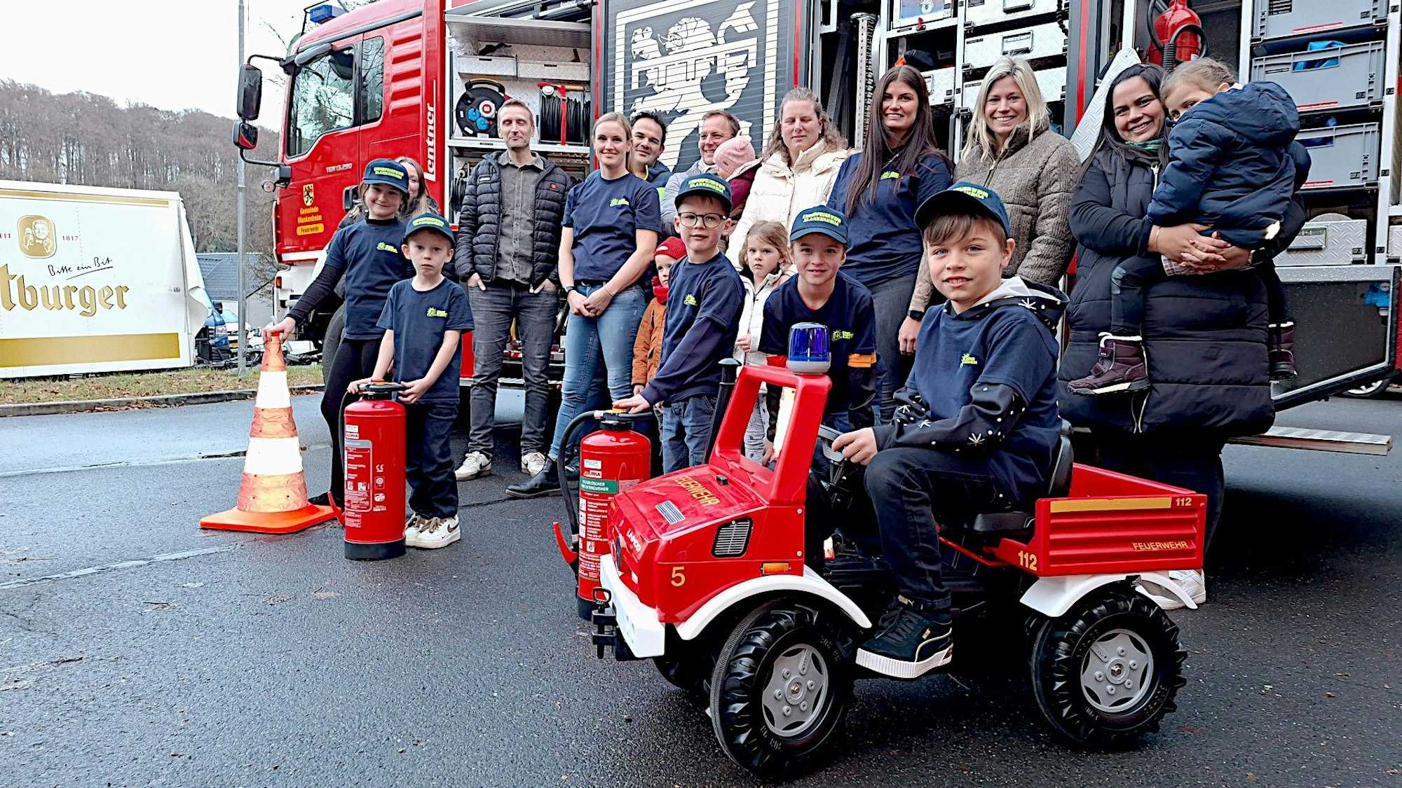 Ein Junge sitzt auf einem Mini-Feuerwehrfahrzeug. Neben ihm stehen weitere Kinder, hinter ihnen einige Erwachsene.