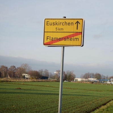 Das Bild zeigt das Flamersheimer Ortsschild, dahinter eine Ackerfläche, rechts eine Straße.