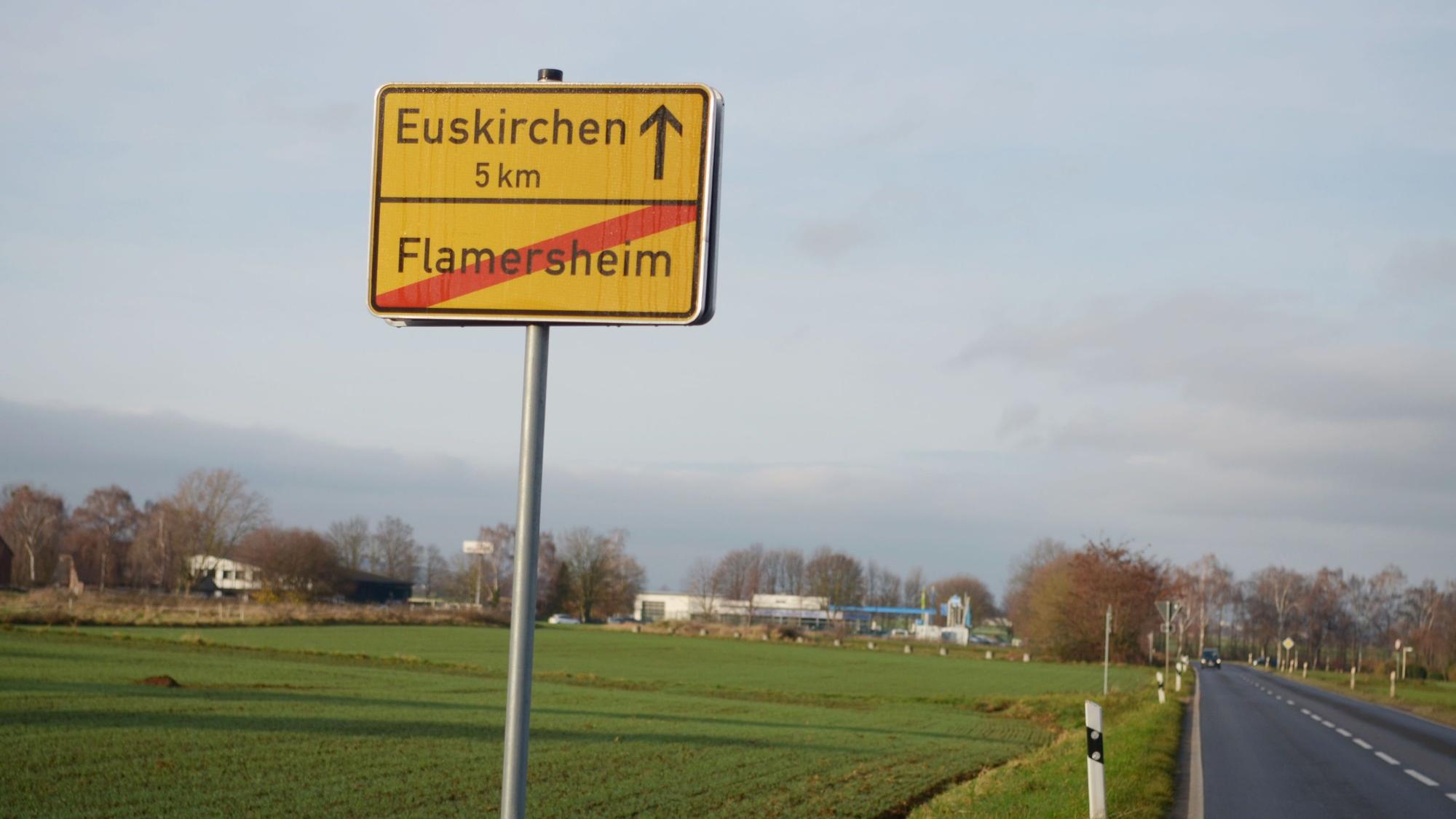 Das Bild zeigt das Flamersheimer Ortsschild, dahinter eine Ackerfläche, rechts eine Straße.