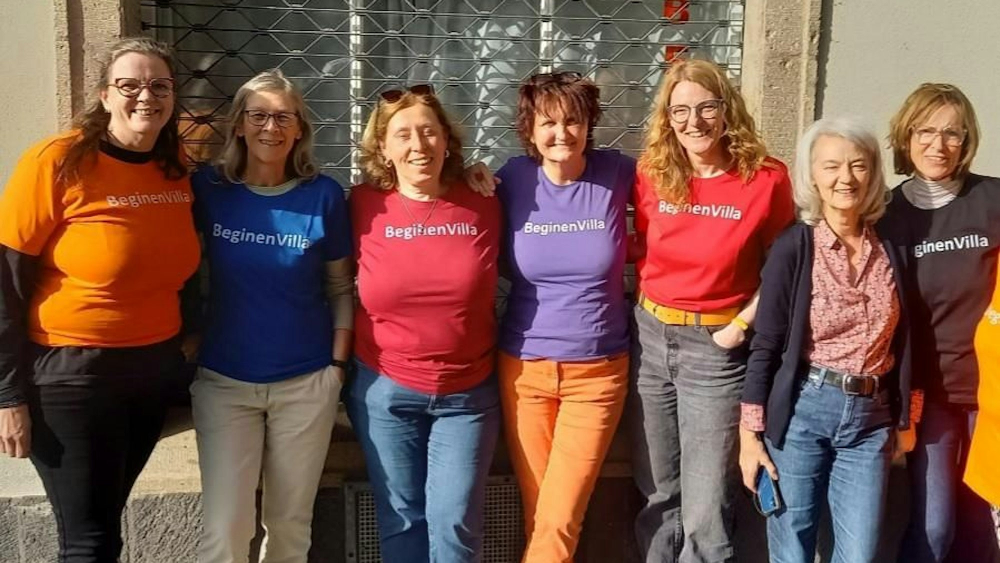 Sieben Frauen stehen Arm in Arm in bunten T-Shirts nebeneinander. Auf denen steht „BeginenVilla“.