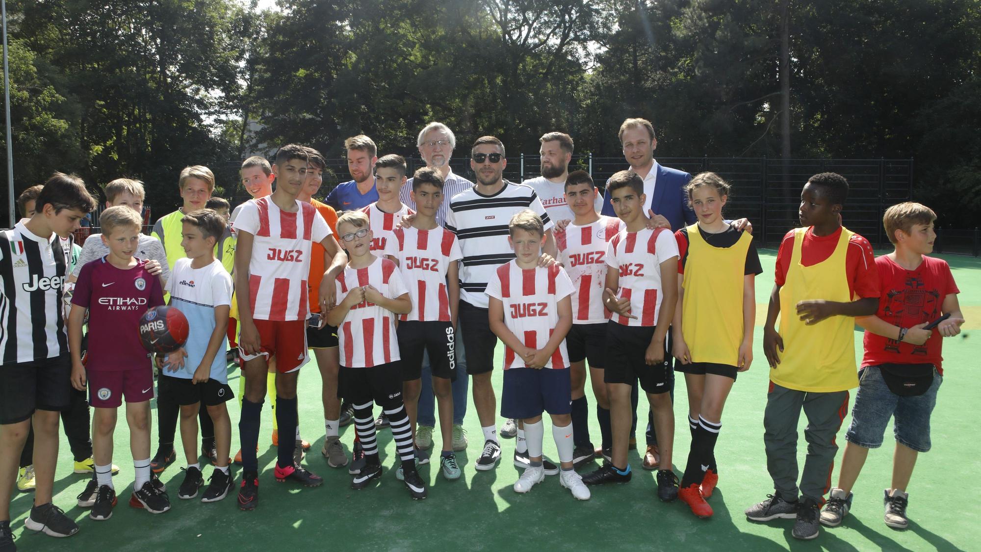 Viele Kinder in Trikots stehen auf einem Bolzplatz mit grünem Belag. In der Mitte ist Lukas Podolski.