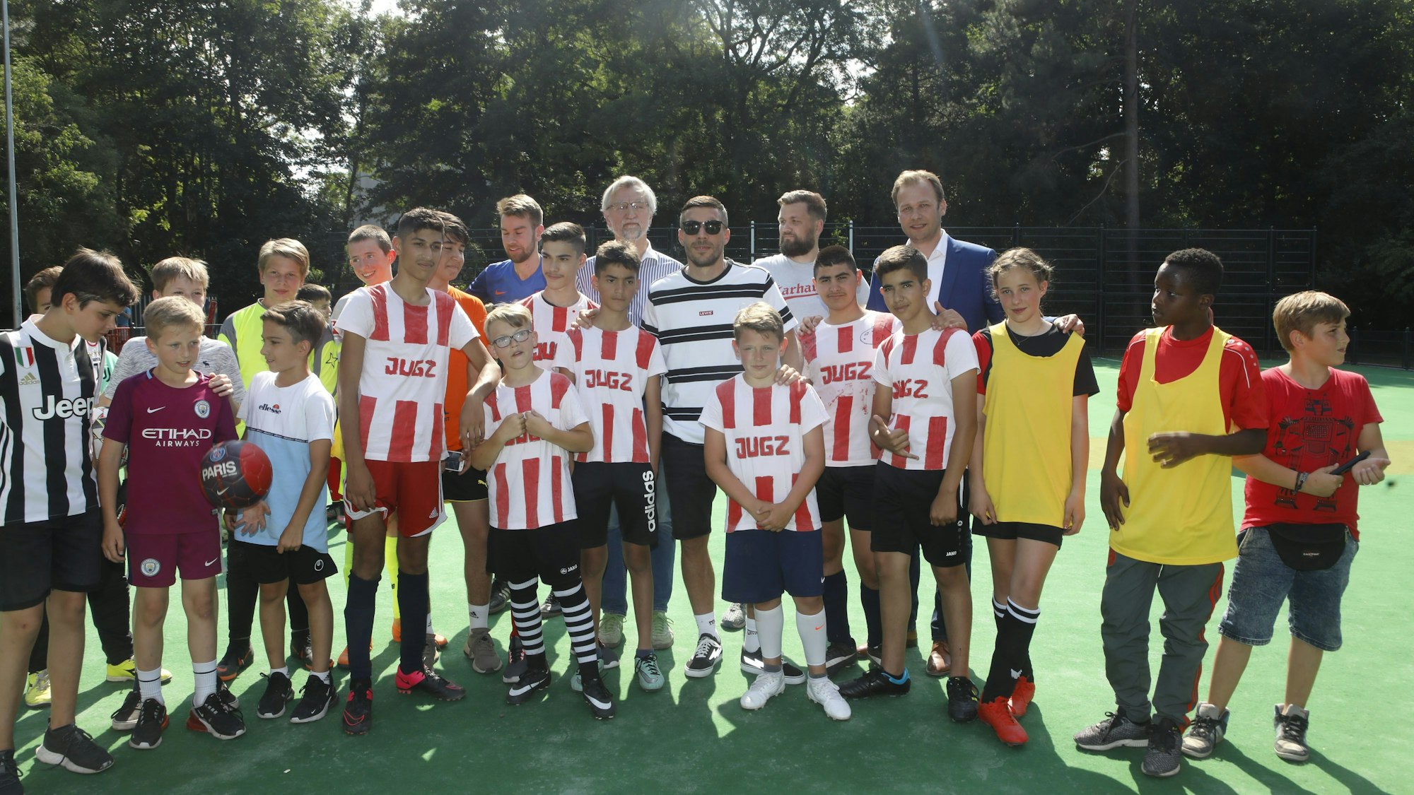 Viele Kinder in Trikots stehen auf einem Bolzplatz mit grünem Belag. In der Mitte ist Lukas Podolski.