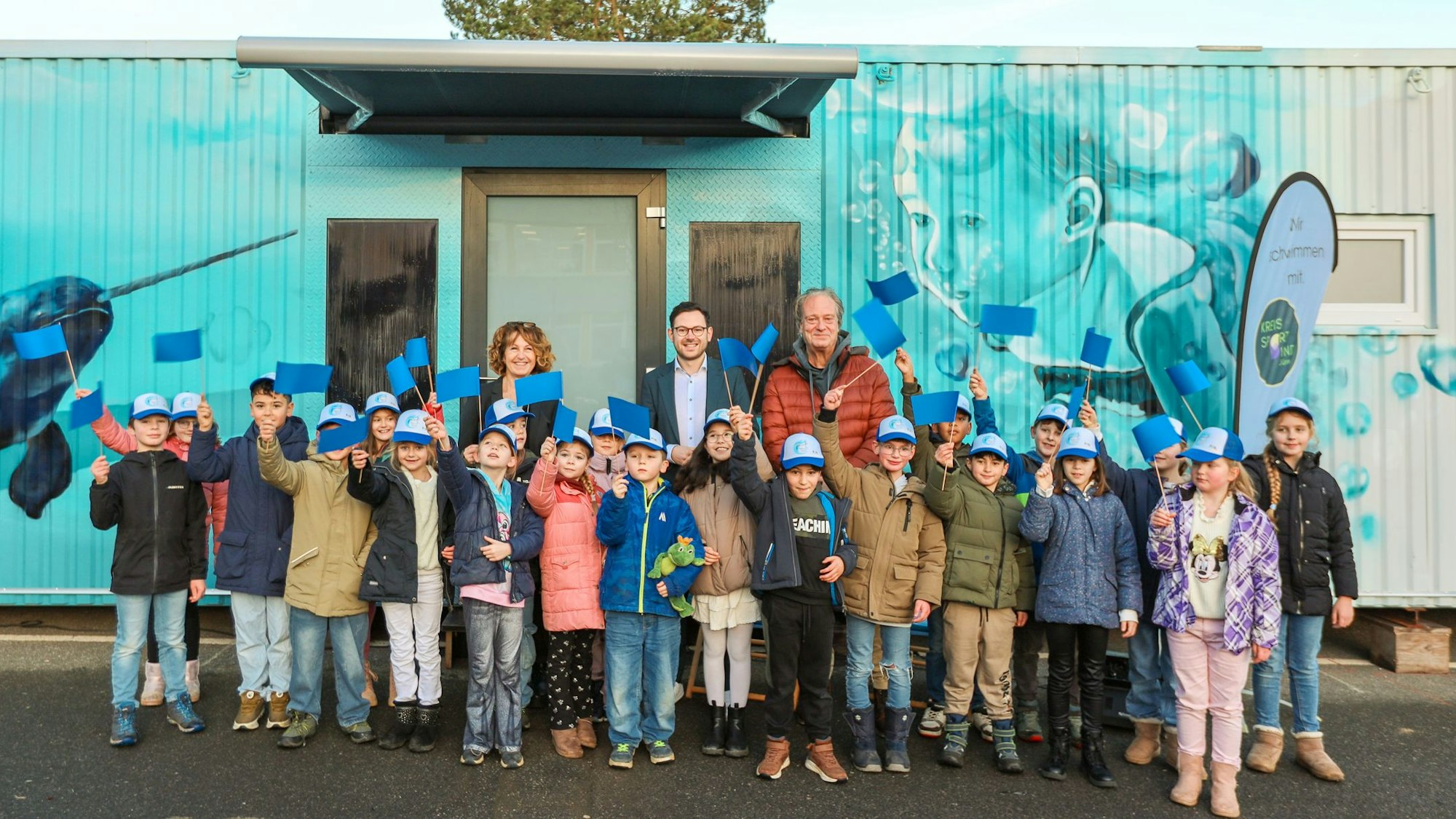 Viele Kinder winken mit blauen Fähnchen, dahinter stehen drei Erwachsene.