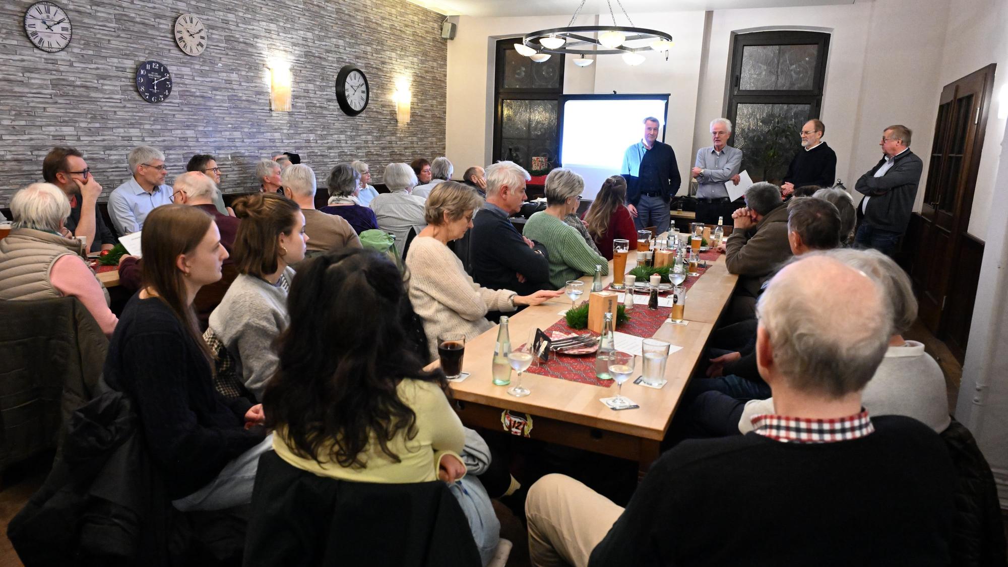 Ein Saal, darin sitzen Männer und Frauen an langen Tischen: vorne stehen vier Männer.