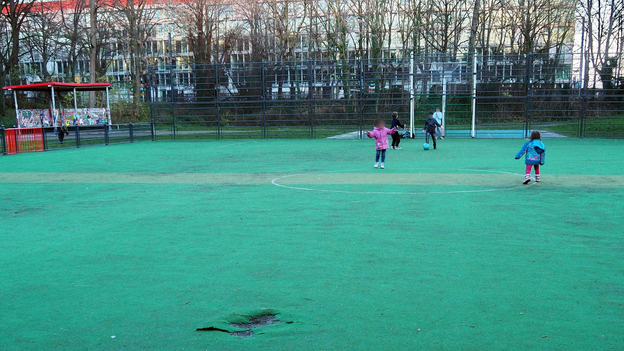 Auf einem mit grünem Bodenbelag ausgestatteten Bolzplatz spielen Kinder Fußball.