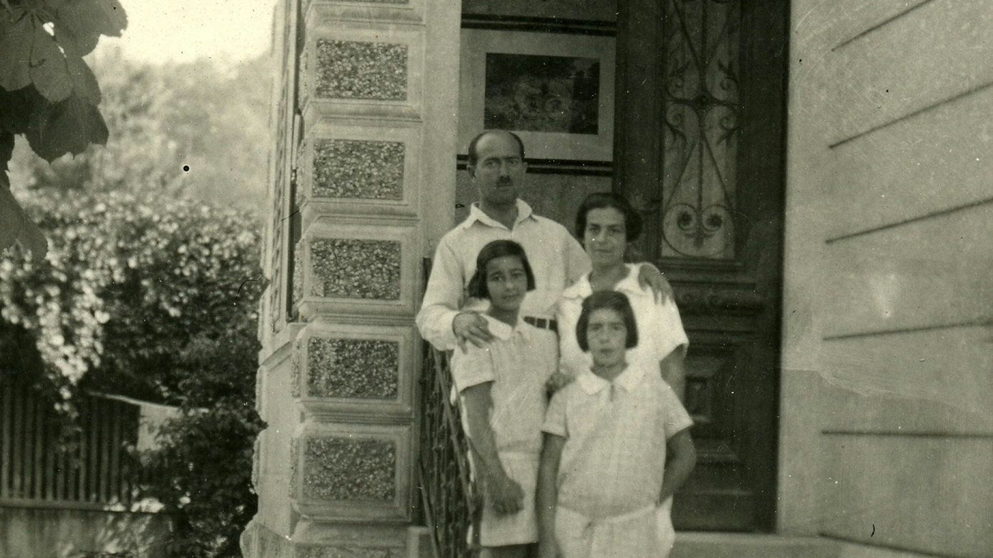 Ein Schwarz-Weiß-Foto zeigt ein Elternpaar mit seinen zwei Töchter auf den Stufen zum Eingang eines großen Hauses.