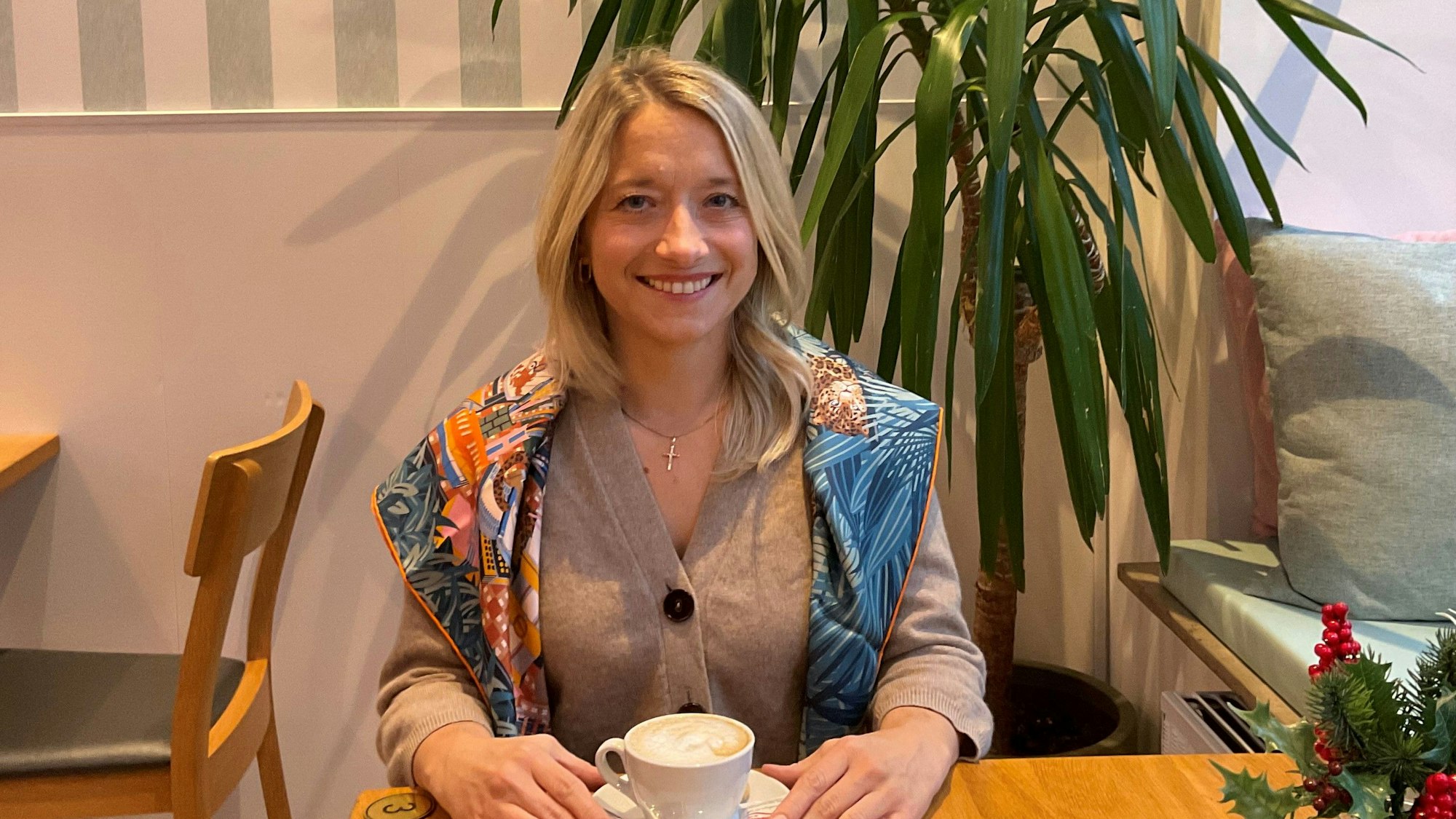 Eine lächelnde Frau sitzt an einem Tisch, mit einem Kaffee vor sich.