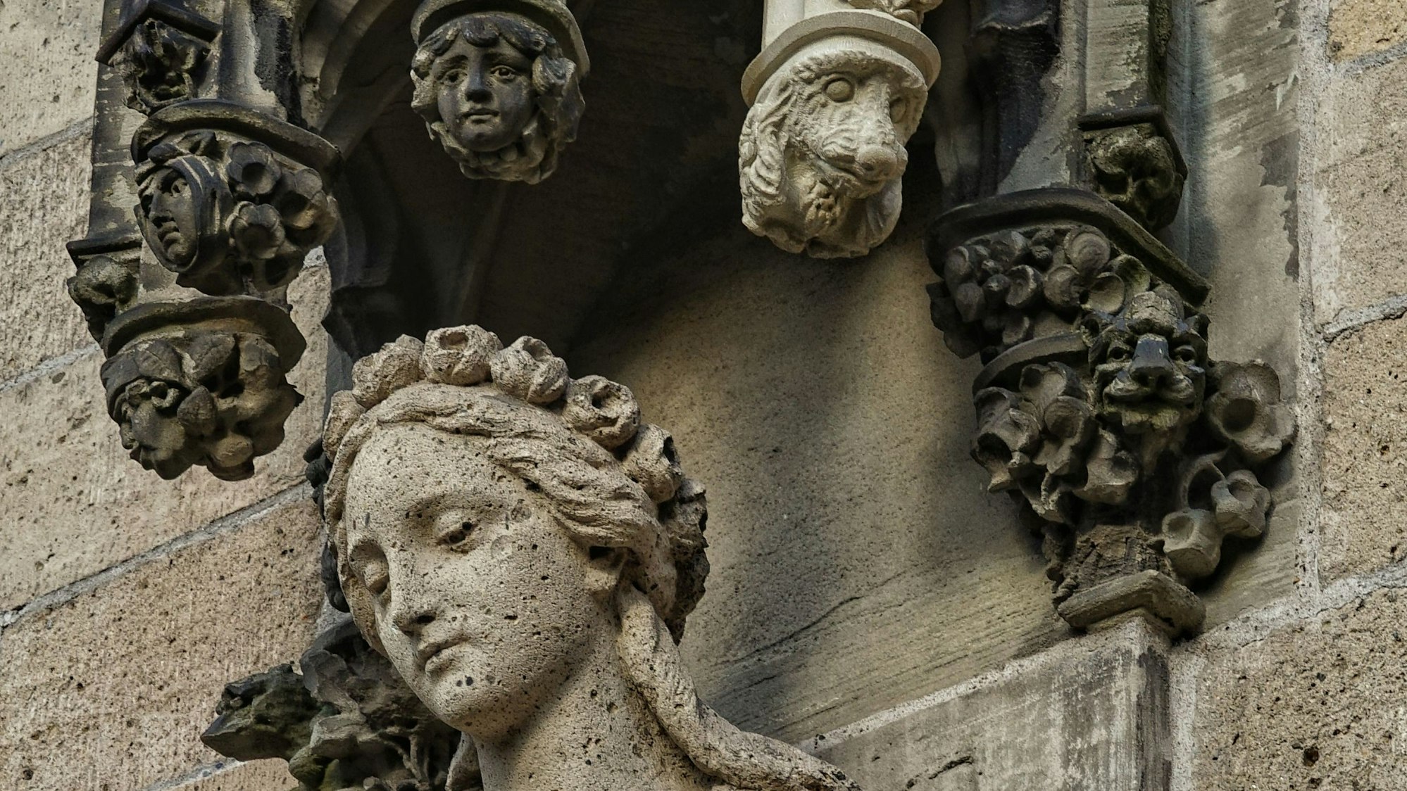 Eine Hundekopf-Skulptur an einem Stein-Baldachin über der Statue einer Frau. An dem Baldachin hänge auch andere Gesichter.