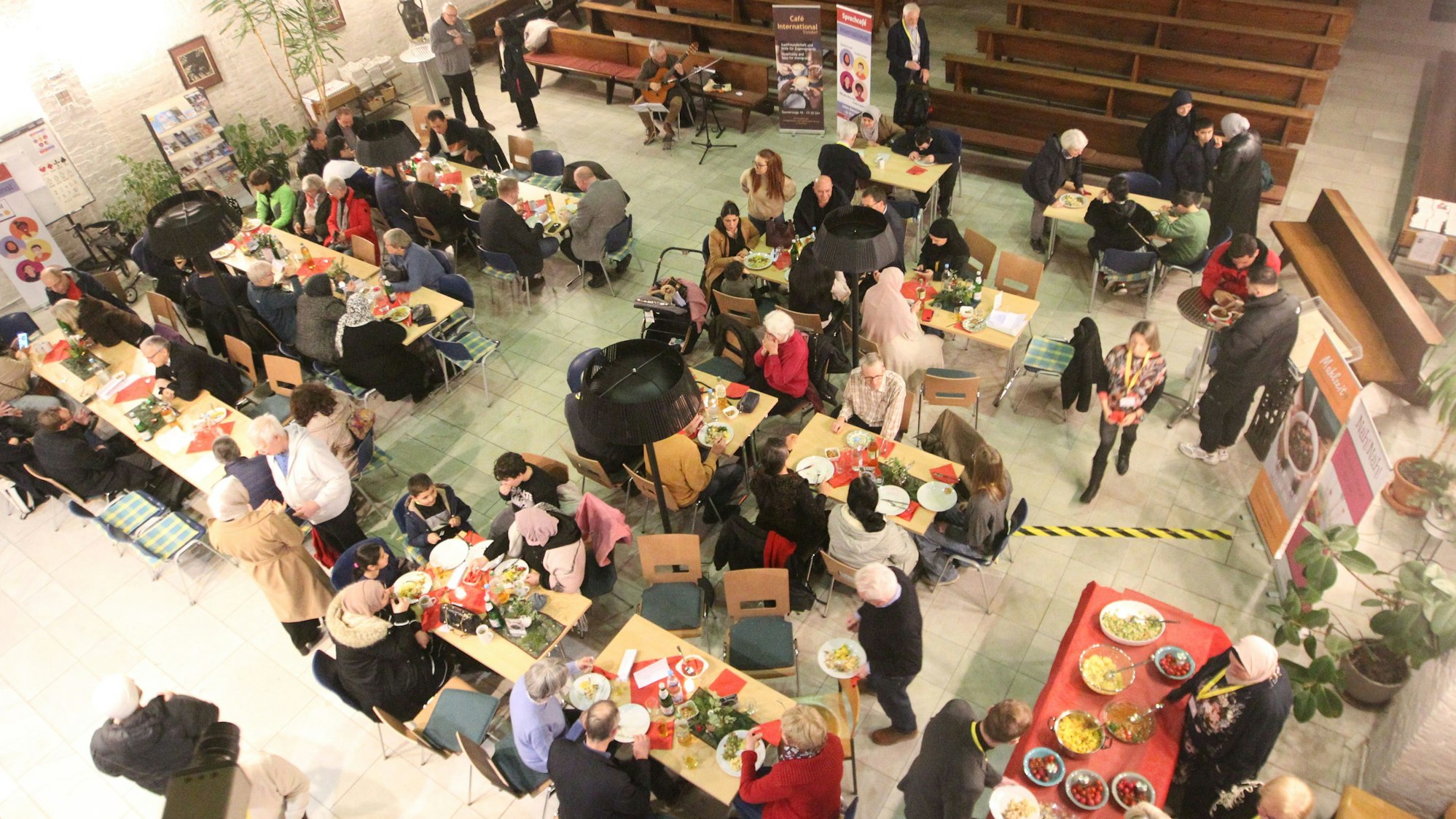Ein Kirchenraum von oben, Menschen sitzen an gedeckten Tischen in dem halb leergeräumten Raum.