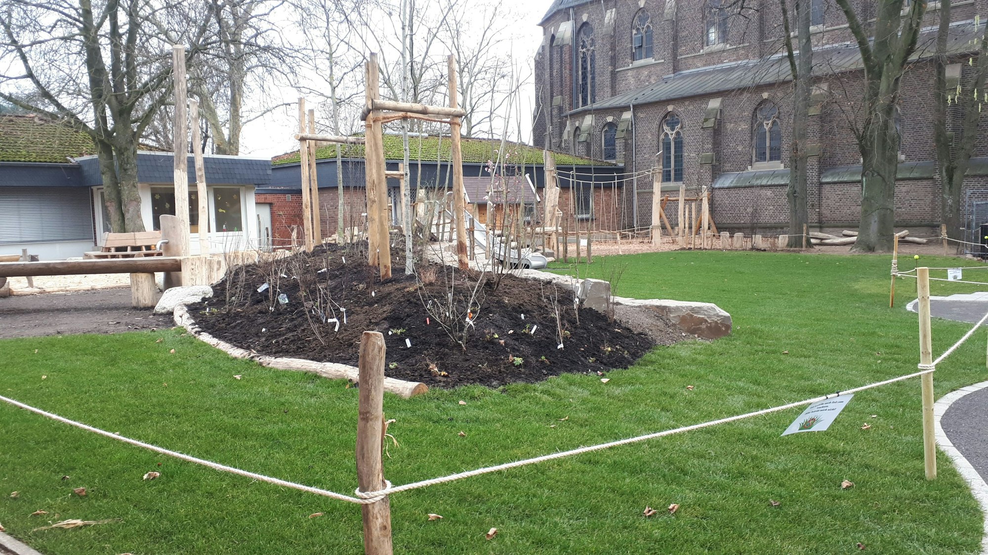 Hinter einer Abzäunung liegen eine Rasenfläche und ein Beethügel mit frisch gepflanzten Sträuchern.