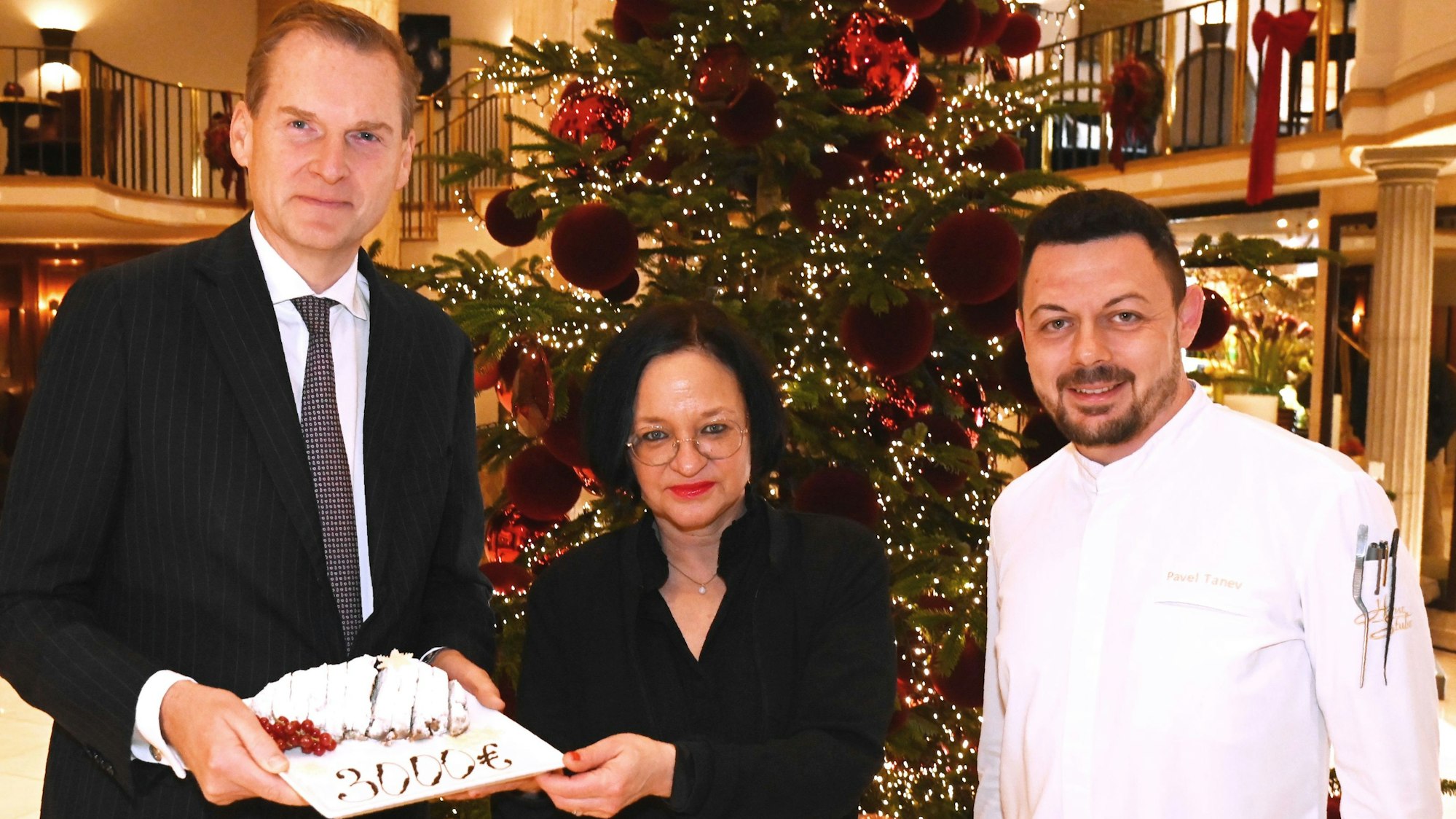 Excelsior Hotel Ernst-Direktor Georg Plesser (li.) und Chef-Patissier Pavel Tanev übergeben die symbolische Spende in Form eines Stollens an wir helfen-Geschäftsführerin Caroline Kron.