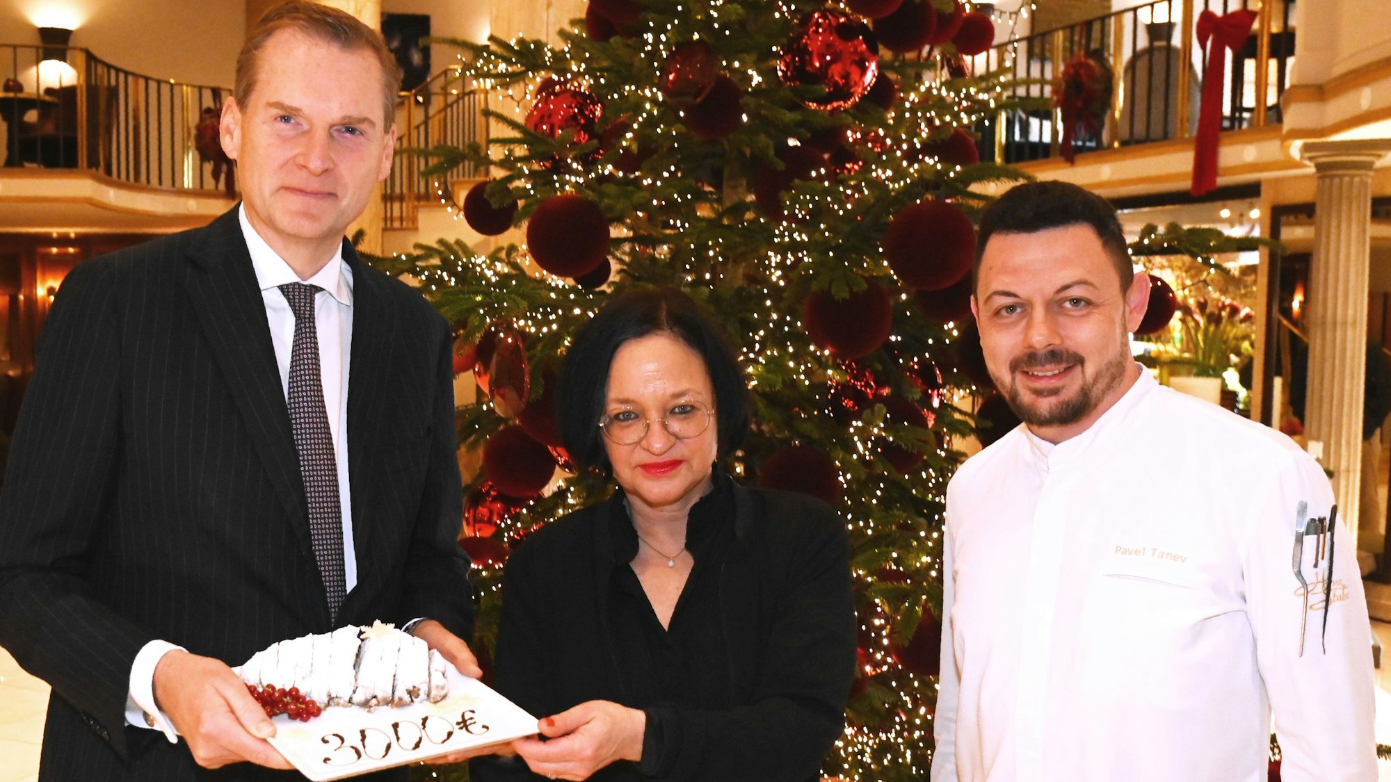 Excelsior Hotel Ernst-Direktor Georg Plesser (li.) und Chef-Patissier Pavel Tanev übergeben die symbolische Spende in Form eines Stollens an wir helfen-Geschäftsführerin Caroline Kron.