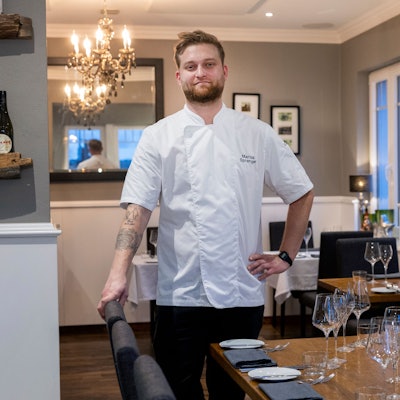 Ein junger Mann steht in weißem Koch-Hemd in seinem Restaurant.